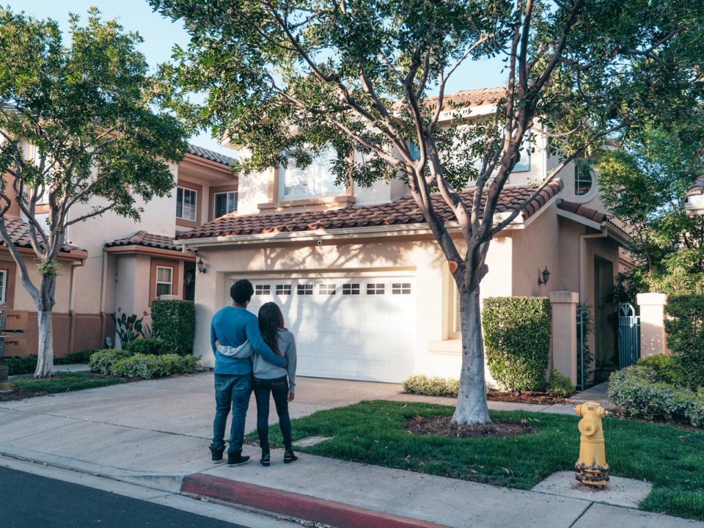 A couple standing together in front of their beautiful home, surrounded by trees and greenery.