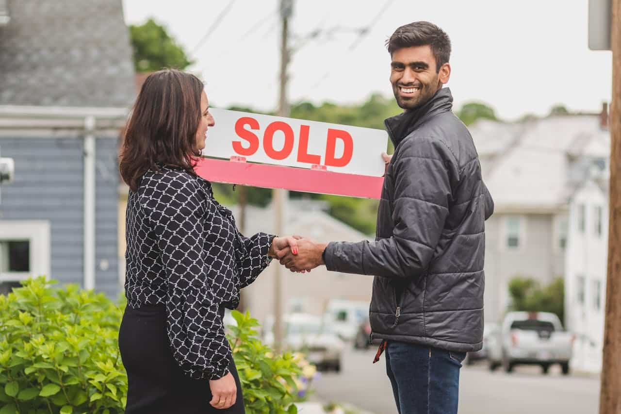 A real estate agent and a happy client shaking hands in front of a sold sign.