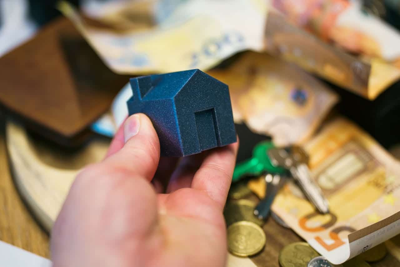 A person holding a miniature house model above a table with money and keys.