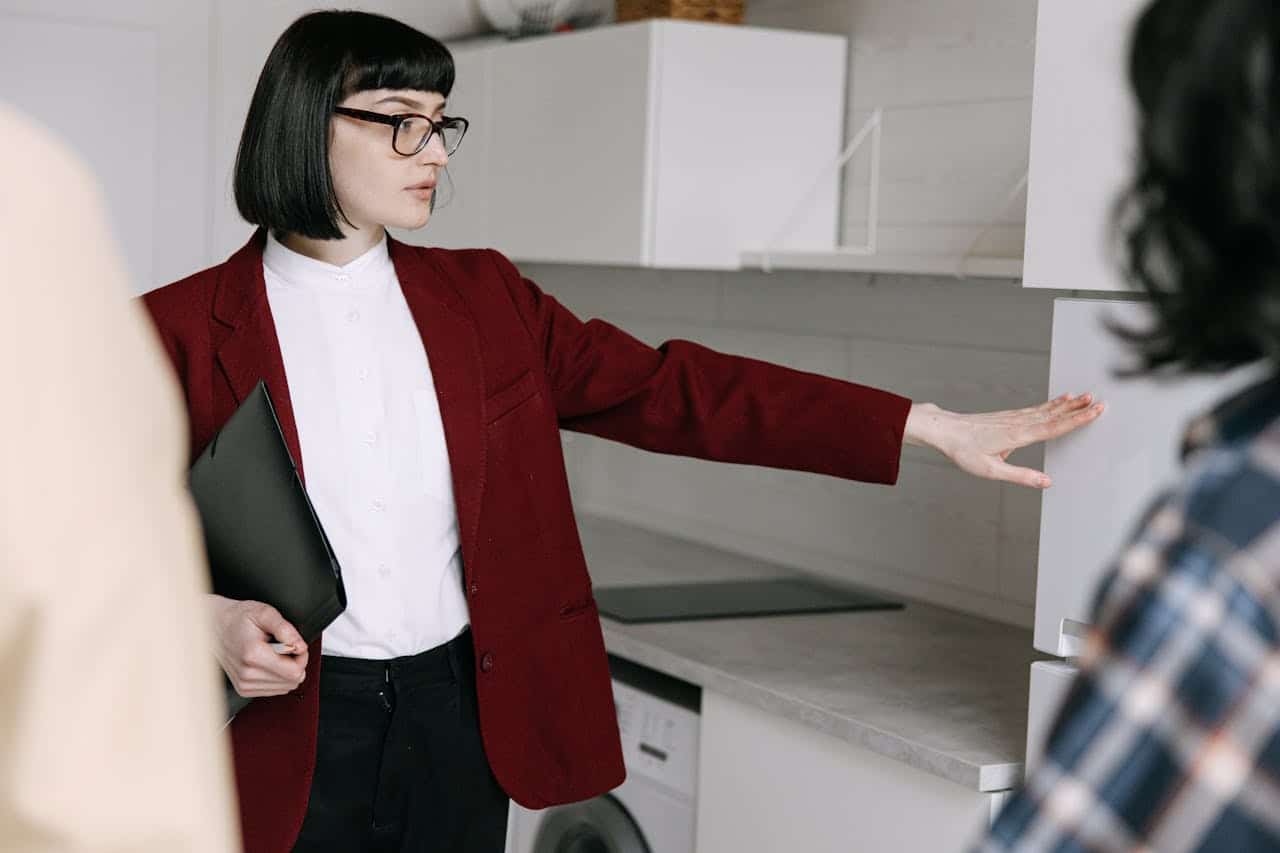 A professional woman in a burgundy blazer explaining kitchen design concepts while pointing at cabinets.
