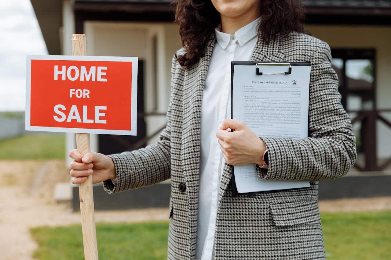 A woman smiling while holding a sign that reads "Home for Sale" in front of a house.