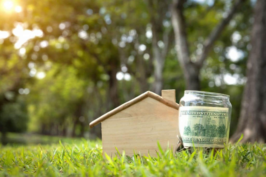 A wooden house model beside a jar filled with money, symbolizing savings and investment.