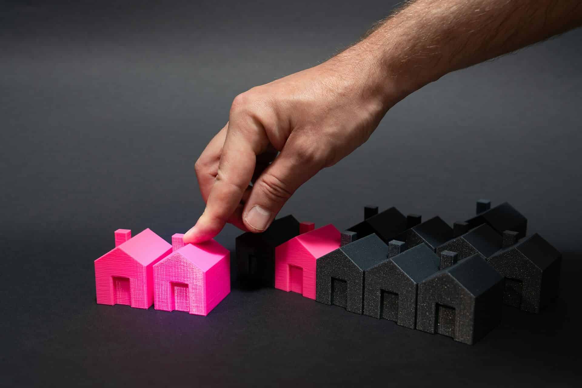A hand selecting a pink house among black houses on a dark background.