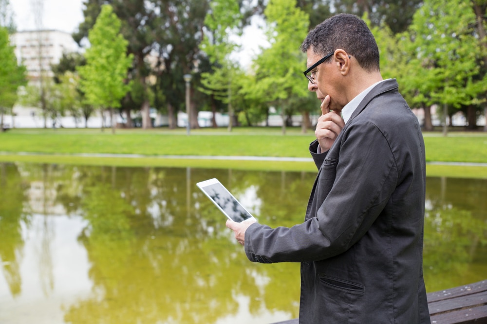 A man thoughtfully using a tablet by a serene pond in a park.