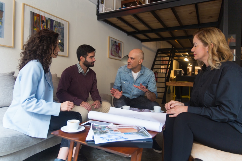 A group of four professionals engaged in a brainstorming session over coffee in a modern office setting.