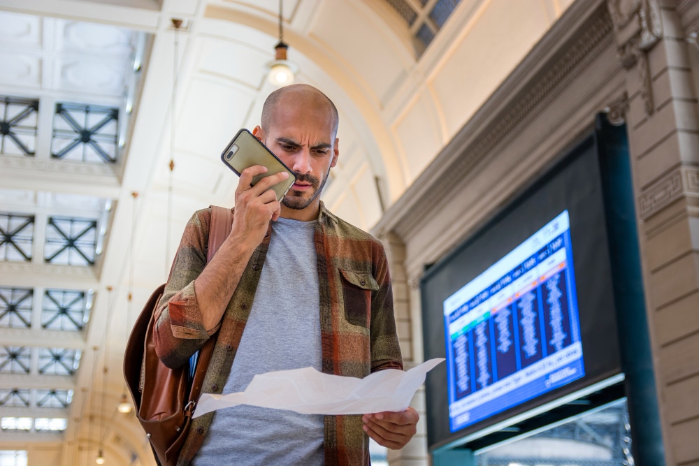 A man looking at a paper while talking on his phone in a train station.
