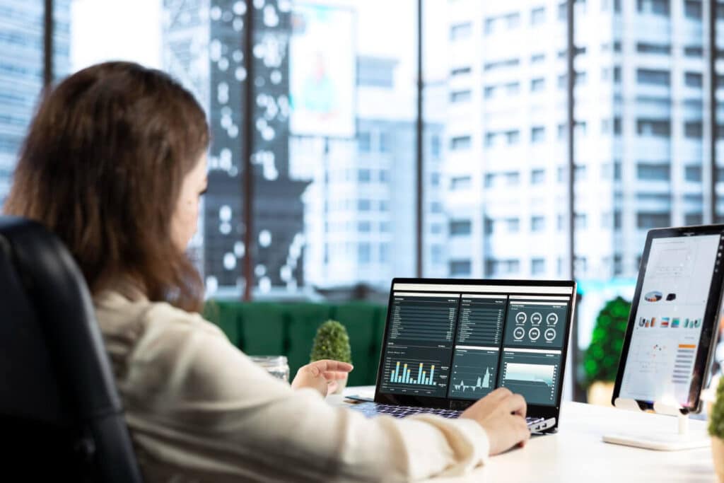 A data analyst reviewing charts and graphs on a laptop in a modern office setting.