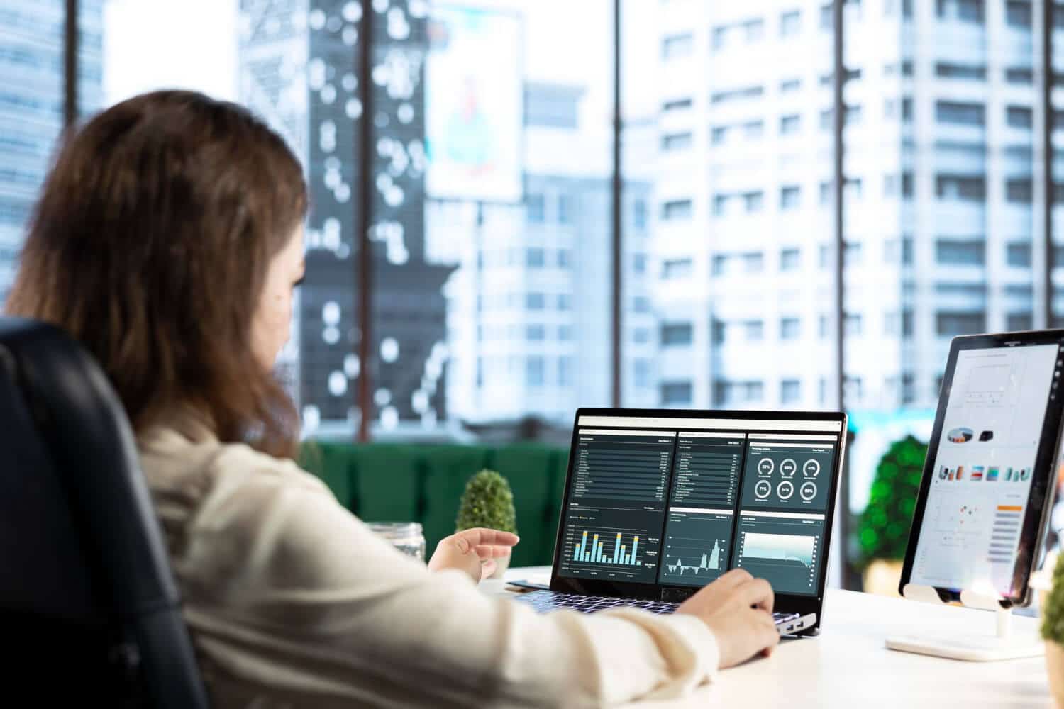 A data analyst reviewing charts and graphs on a laptop in a modern office setting.
