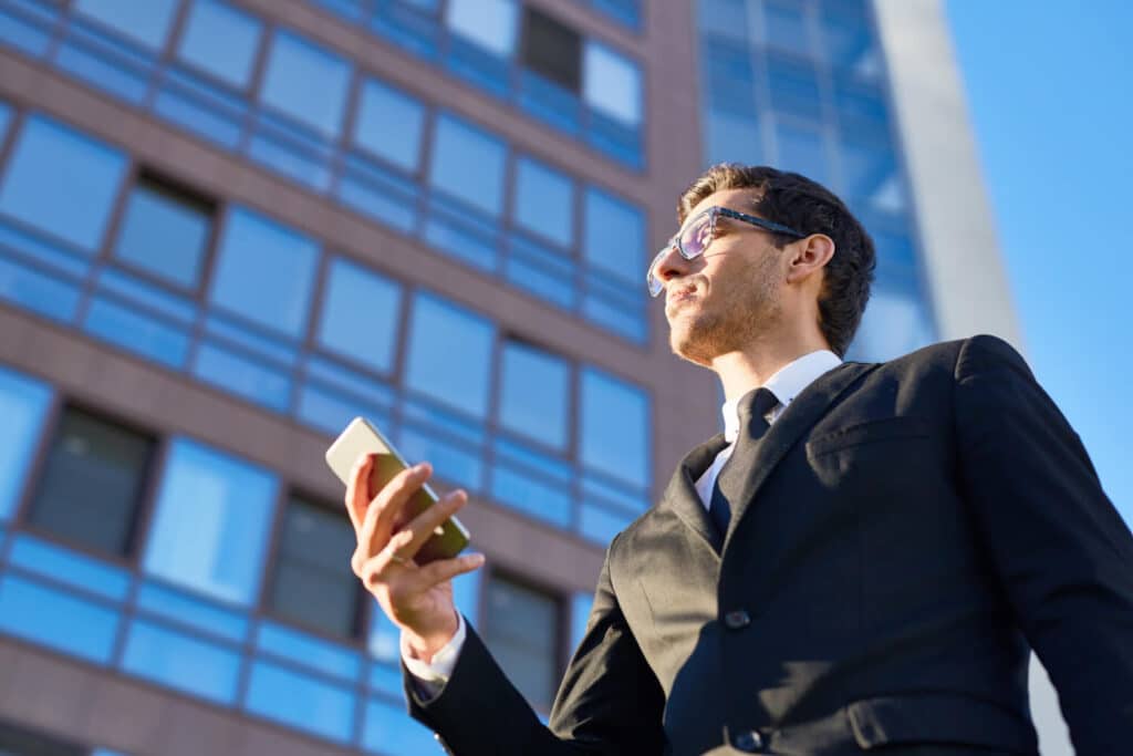 A professional man in a suit holding a smartphone while looking up at a modern building.